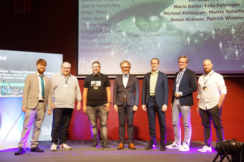 Ein Gruppenbild der Vortragenden der SPS-Tagung. Im Hintergrund befinden sich eine Werbetafel der HOK und eine Leinwand mit einer Präsentation.