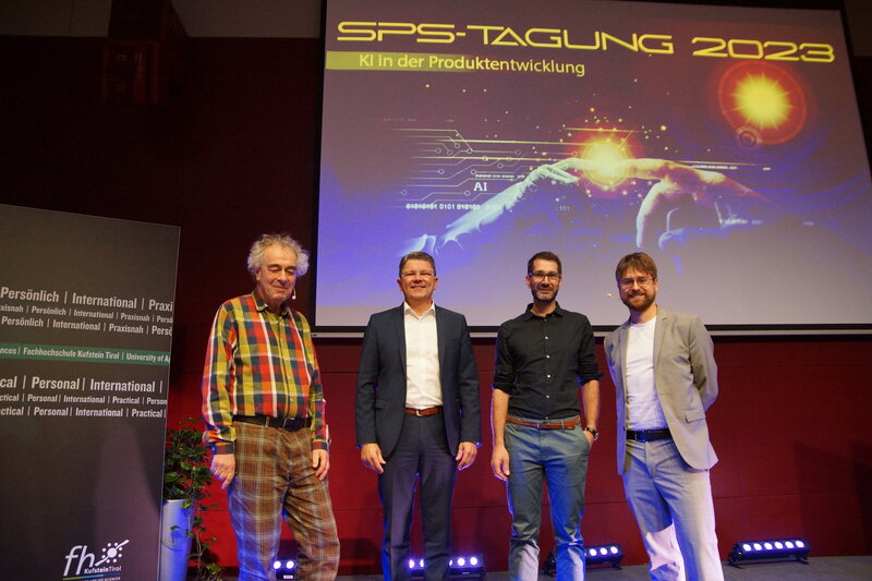 Vier Vortragende der SPS-Tagung stehen auf der Bühne und machen ein Gruppenfoto. Hinter ihnen befindet sich eine Leinwand mit einer PowerPoint-Präsentation.