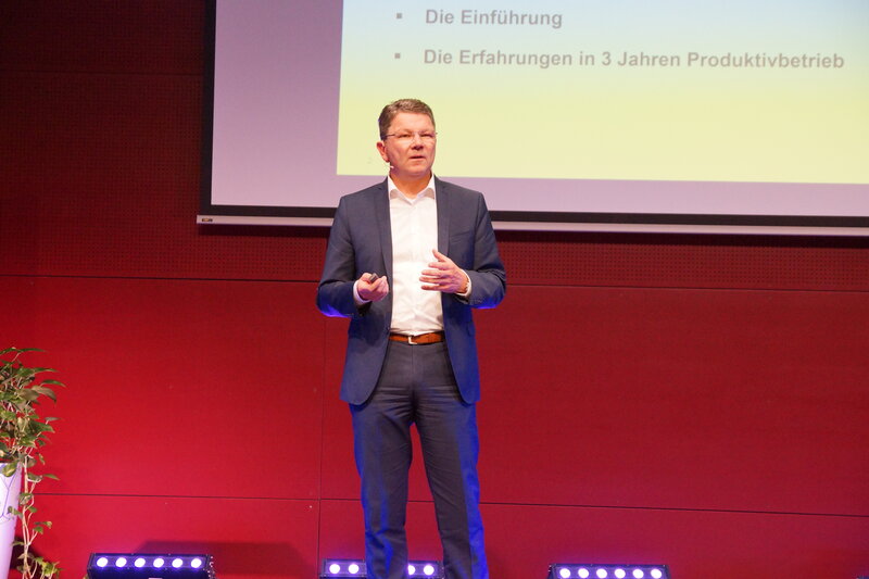 Ein Vortragender steht auf der Bühne. Hinter ihm befindet sich eine große Leinwand, auf der eine PowerPoint-Präsentation angezeigt wird.