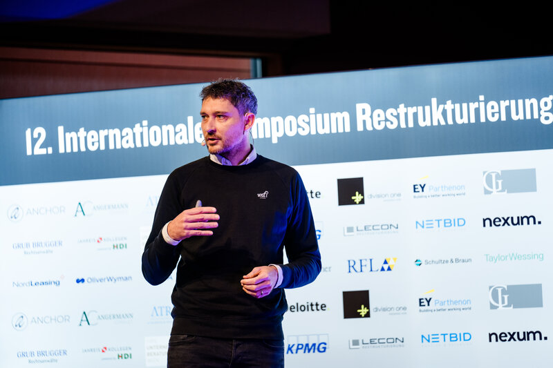 Christian Lammers hält eine Präsentation beim 12. Symposium Restrukturierung der HOK. Hinter ihm befindet sich eine Wand mit Sponsoring-Logos.
