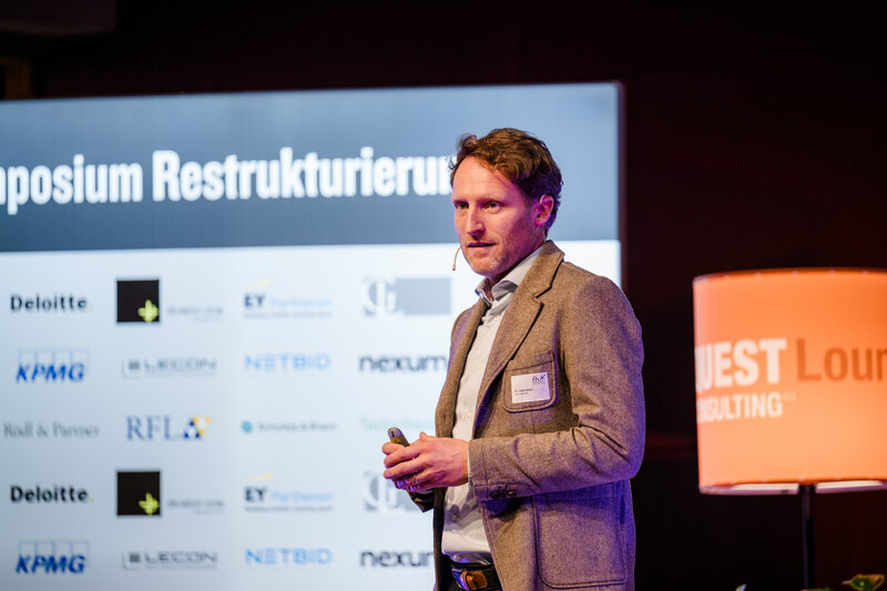 Dr. Lothar Stadler hält eine Präsentation im Veranstaltungsraum im Rahmen des 12. Internationalen Symposium Restrukturierung der HOK. Hinter ihm befindet sich eine Wand mit Sponsoring-Logos.