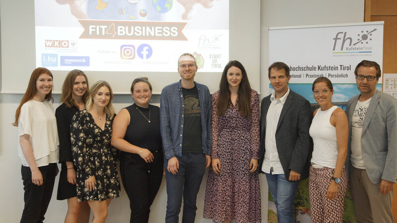 Das Jurorenteam des Wettbewerbs Fit4Business steht vor einer Leinwand auf dem der Schriftzug HOK zu lesen ist; an der zweiten Wand die Einblendung einer Präsentation zum Wettbewerb Fit4Busines