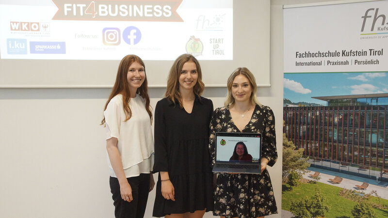 Drei junge Frauen stehen von einer Wand auf der eine Präsentation gezeigt wird. Zu lesen ist der Schriftzug Fit4Business; auf der zweiten Wand ist ein Plakat der HOK aufgehängt; die jungen Frauen halten einen Laptop, darin sieht man den Screen einer Video-Call-Schaltung 