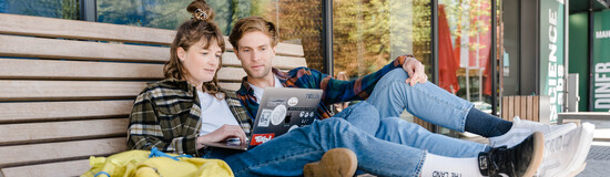 Zwei Studierende arbeiten gemeinsam am Laptop auf einer Bank vor der HOK
