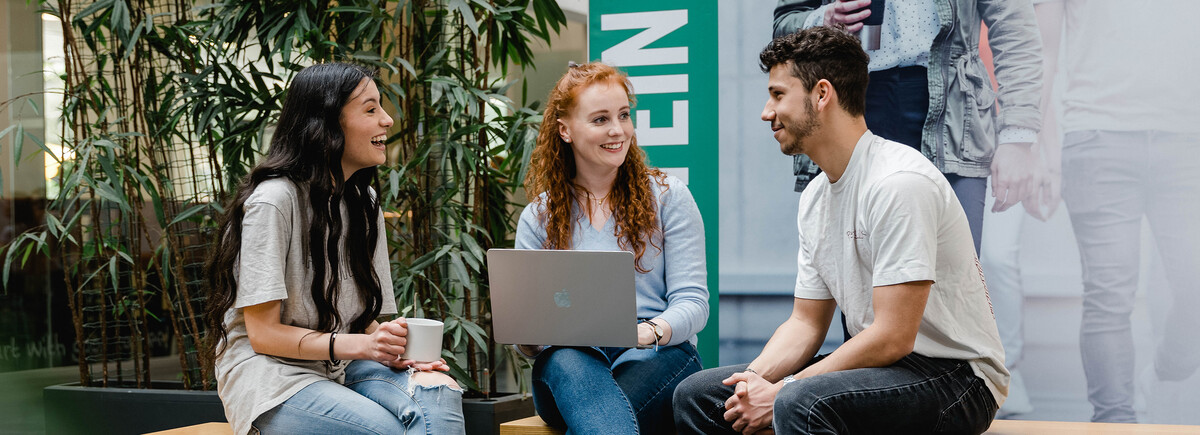 Drei Studierende der HOK sitzen zusammen in der Aula, unterhalten sich und arbeiten am Laptop.