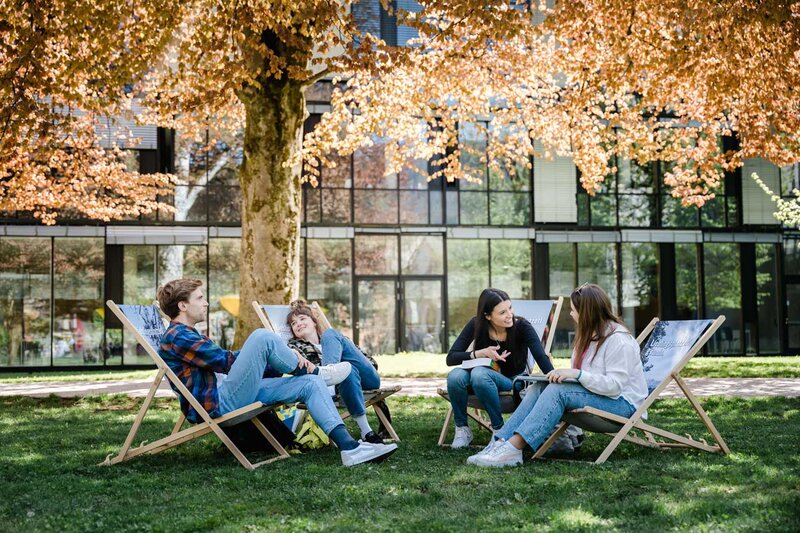 Vier Studierende der HOK sitzen im Park vor dem Campus in Liegestühlen und unterhalten sich bei sonnigem Wetter.