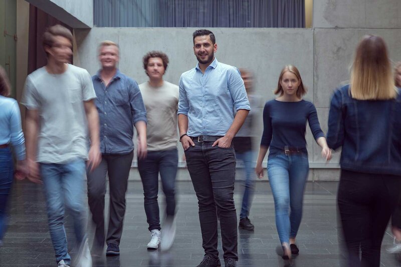 Viele Personen bewegen sich durch die Aula der HOK, und im Zentrum des Bildes steht ein Mann und blickt in die Kamera.