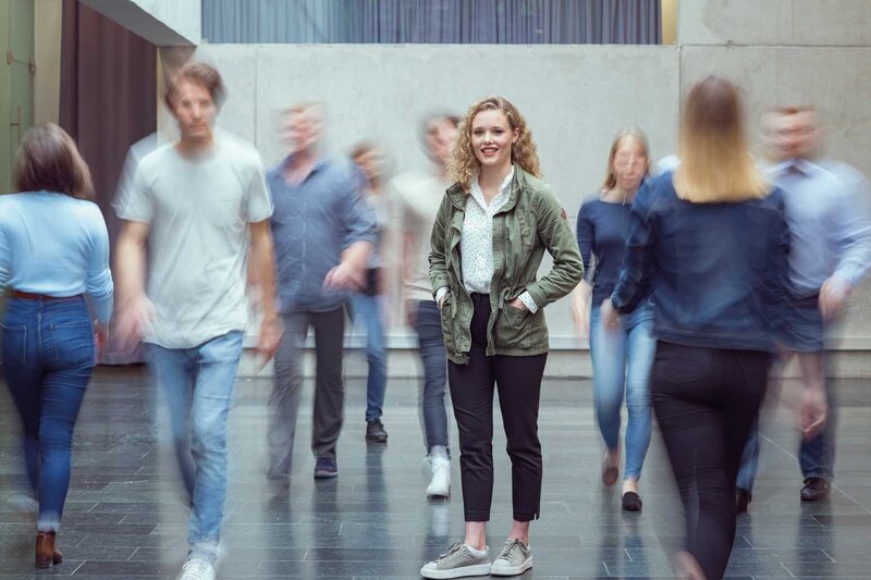Viele Personen bewegen sich durch die Aula der HOK, und im Zentrum des Bildes steht eine Frau und blickt in die Kamera.
