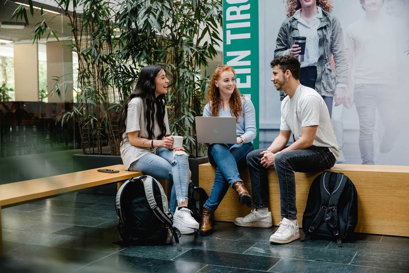 Drei Studierende der HOK sitzen zusammen in der Aula, unterhalten sich und arbeiten am Laptop.