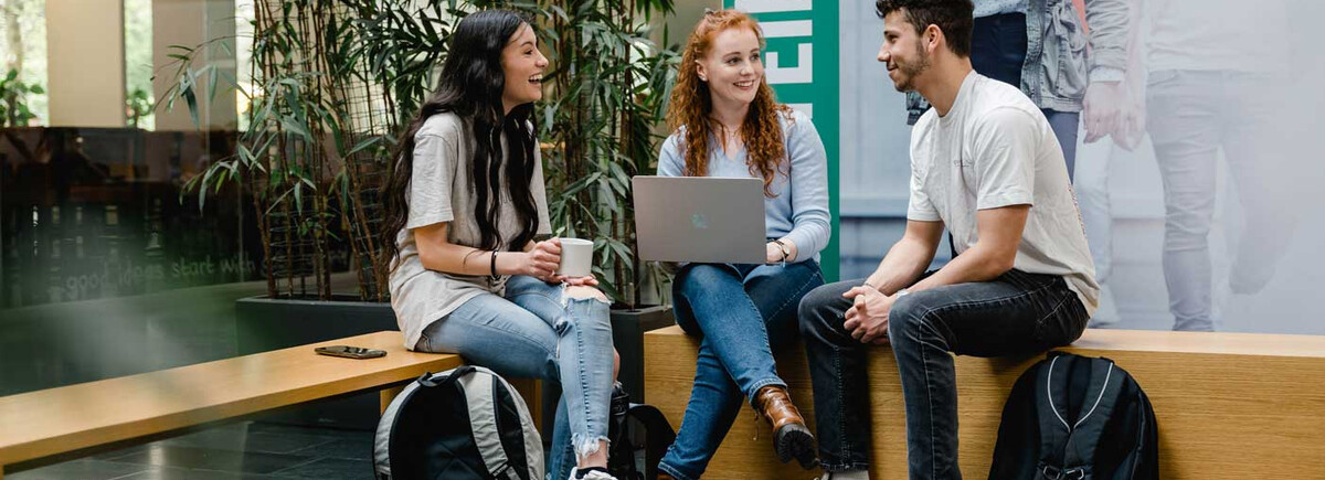 Drei Studierende der HOK sitzen zusammen in der Aula, unterhalten sich und arbeiten am Laptop.