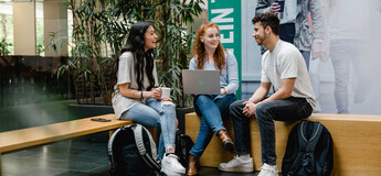 Drei Studierende der HOK sitzen zusammen in der Aula, unterhalten sich und arbeiten am Laptop.