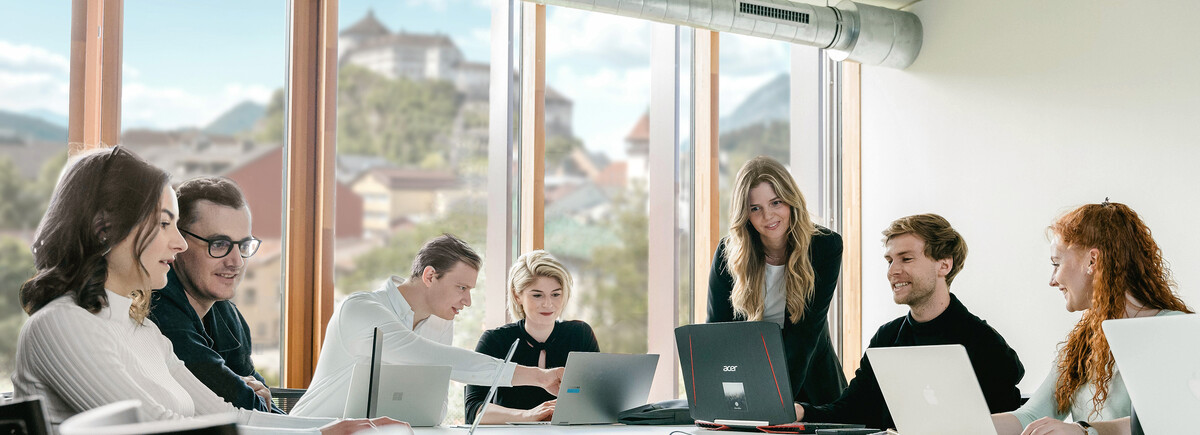 Studierende an der HOK sitzen an einem Tisch mit Laptops und diskutieren in einem hellen Raum mit Blick auf die Festung Kufstein.
