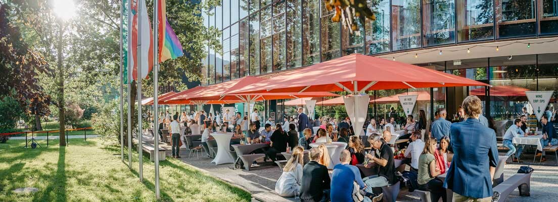 Gäste des Home Coming 2024 der HOK bei sommerlichem Wetter auf der Terrasse des Campus im Gespräch und beim Netzwerken.