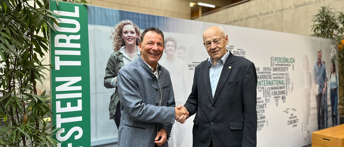 Gründungsvater der HOK Walter J. Mayr zusammen mit Geschäftsführer Thomas Madritsch vor einem Banner in der Aula der Fachhochschule.
