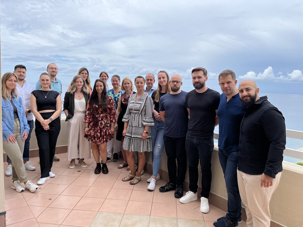 Eine Gruppe von Personen steht in sommerlicher Kleidung auf einer Dachterrasse, im Hintergrund das Meer.