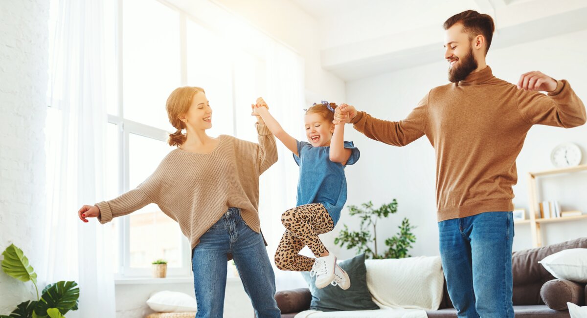 röhliche Eltern schwingen ihr Kind zwischen sich, während sie in einem sonnendurchfluteten Wohnzimmer stehen. Alle tragen lässige Freizeitkleidung und genießen den Moment gemeinsam.