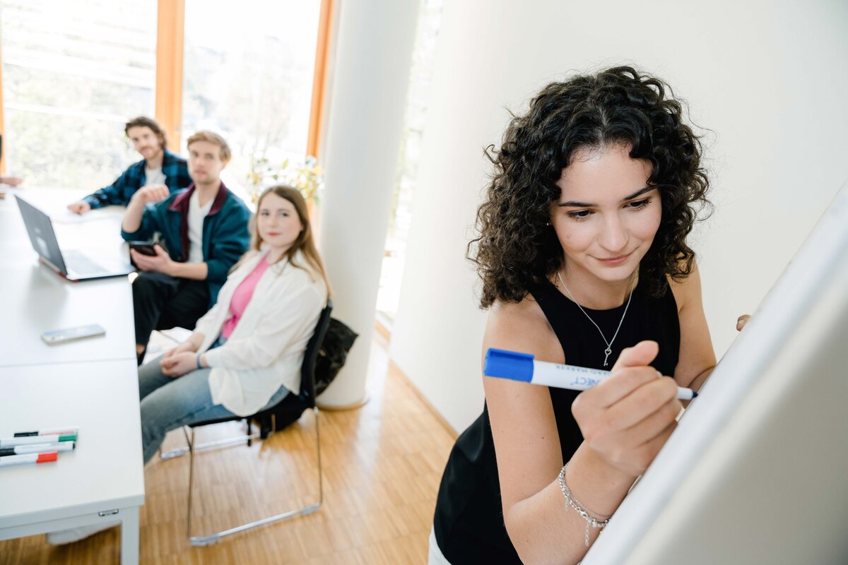 Junge Frau schreibt mit einem blauen Marker auf ein Whiteboard, während drei Studierende ihr von einem Tisch aus zuschauen.