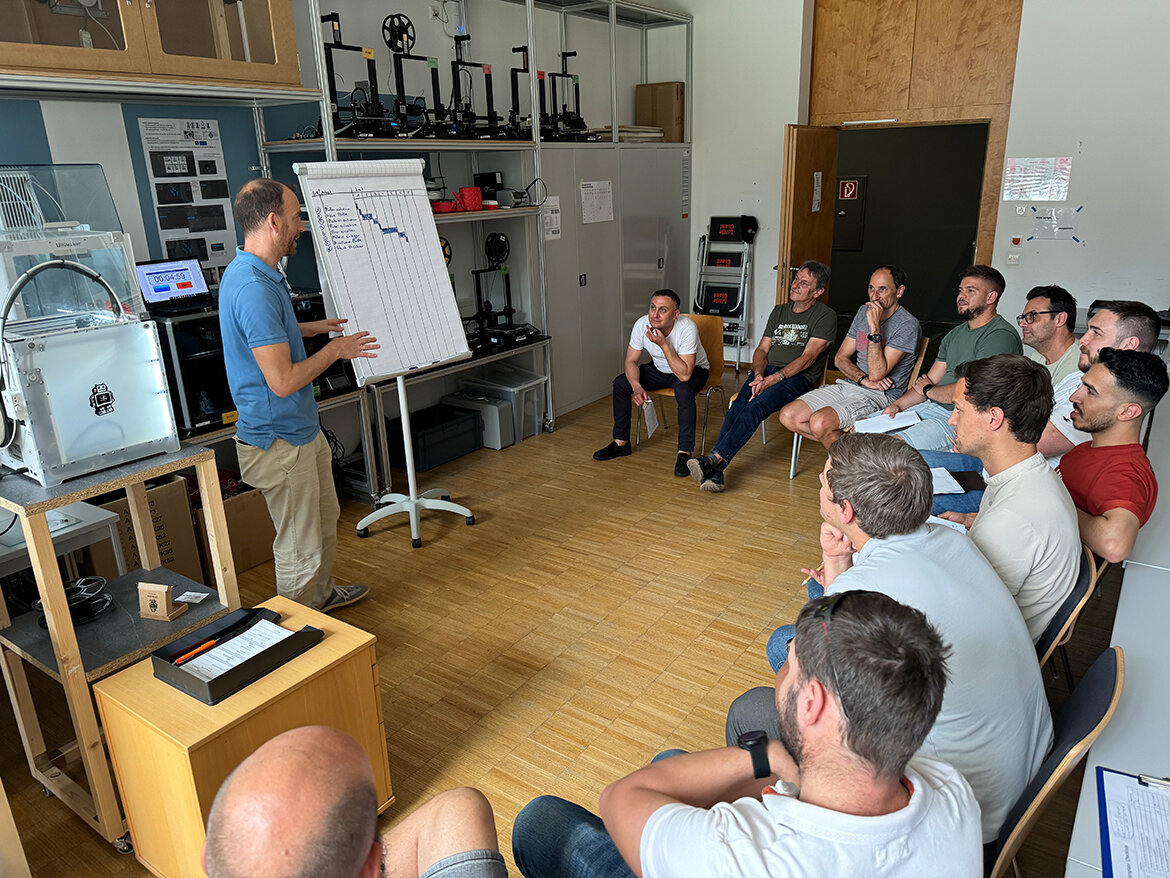 Männer sitzen auf Stühlen in einem Kreis und schauen zu einem Mann im blauen Polo-Hemd, der vor einem Flipchart steht.