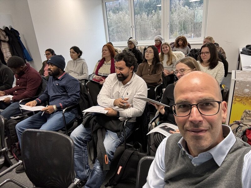 Ein Mann mit Glatze sitz vor einer Gruppe von Menschen auf einem Stuhl und macht ein Selfie.