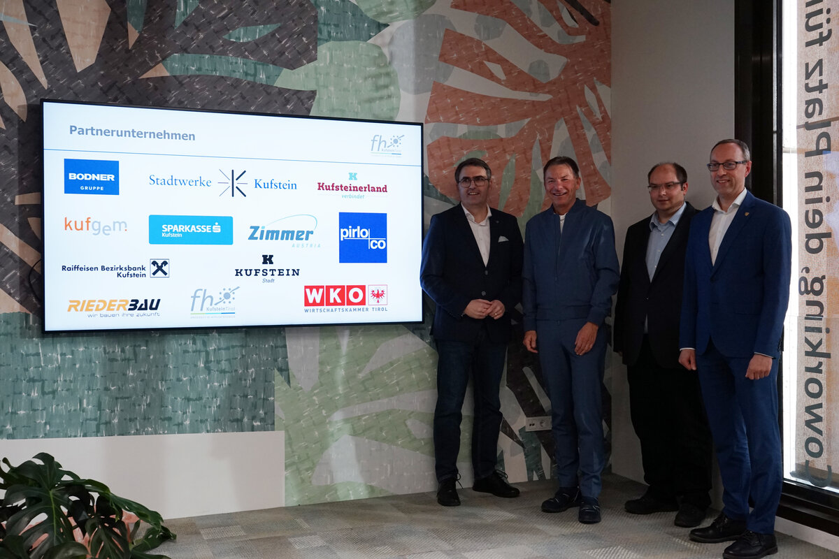 Vier Männer in Anzügen stehen rechts neben einem Bildschirm mit Logos.