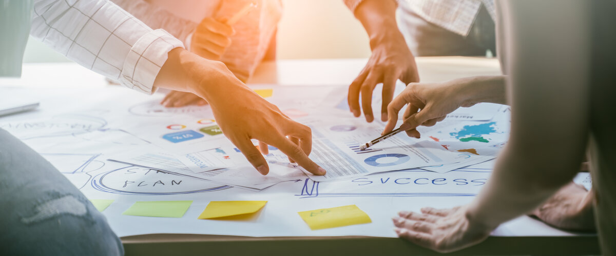 Hände auf einem Arbeitstisch während eines Meetings, mit sichtbaren Post-Its und einem Marketing-Plan im Hintergrund.
