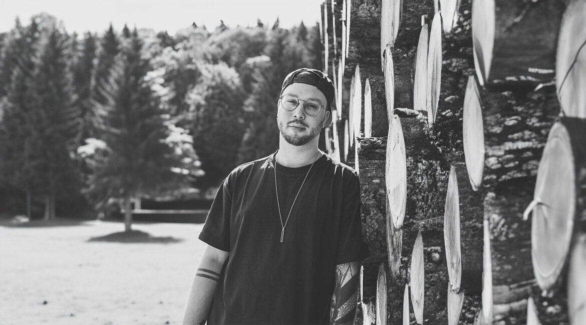 Tobias lehnt an einem Stapel Holzstämme und blickt in die Kamera. Im Hintergrund sind Bäume und eine Wiese zu sehen.