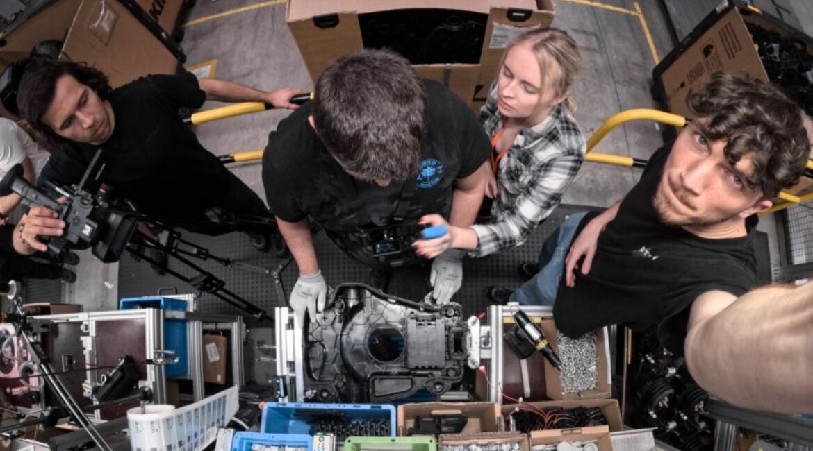 Studierende der HOK und ein Kameraprofi des eLearning Centers richten gemeinsam mit einem Produktionsmitarbeiter von STIHL Tirol die Kamera für Prozessaufnahmen an einer Mähroboter-Montagelinie ein.