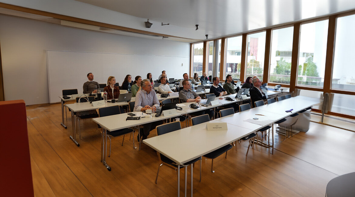 Workshop-Teilnehmer:innen sitzen an Tischen und folgen der Präsentation. Einige machen sich Notizen oder arbeiten an Laptops.