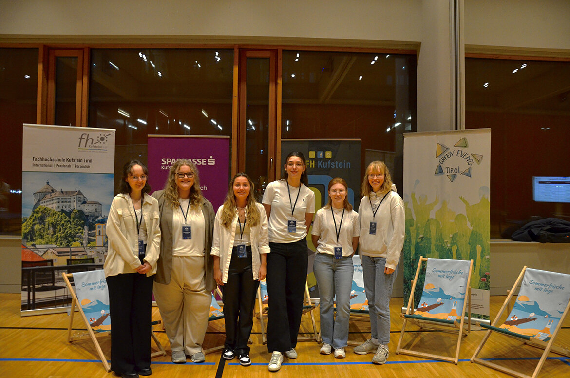 Eine Gruppe von sechs jungen Frauen steht in einer Sporthalle vor Bannern der HOK, der Sparkasse Kufstein und des Green Event Tirol. Sie tragen Namensschilder und lächeln in die Kamera.