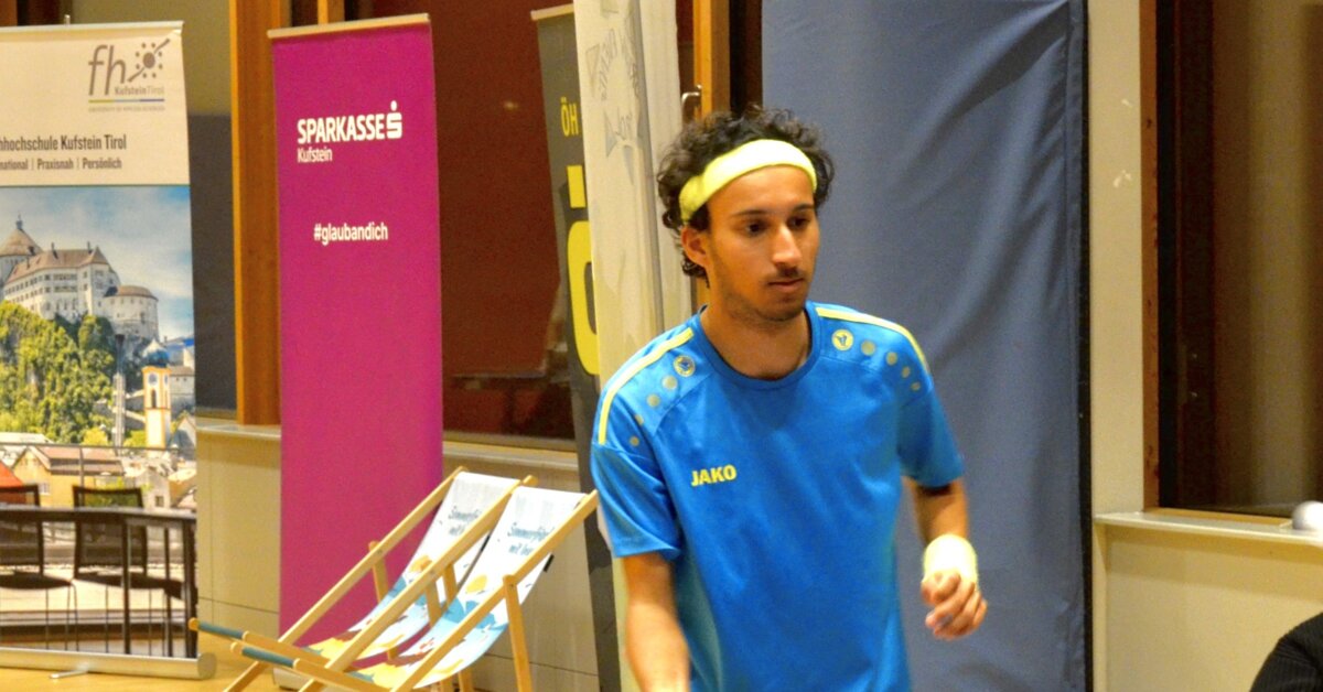Ein Spieler in blau-gelbem Trikot mit Stirnband konzentriert sich während eines Tischtennismatches an der HOK. Im Hintergrund sind Sponsorenbanner und Liegestühle zu sehen.