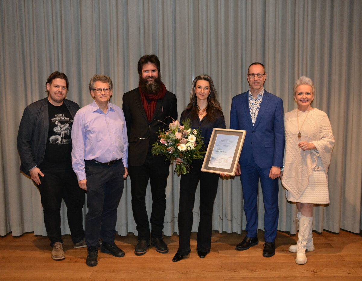Sechs Personen stehen nebeneinander vor einem grauen Vorhang. In der Mitte hält Preisträgerin Zsófia Németh eine Urkunde und einen Blumenstrauß. Neben ihr stehen Vertreter aus Bildung, Kultur und Stadtpolitik, darunter Harald Stoiber, Reinhold Weichselbraun, Klaus Reitberger, FH-Rektor Mario Döller und Vizebürgermeisterin Brigitta Klein.