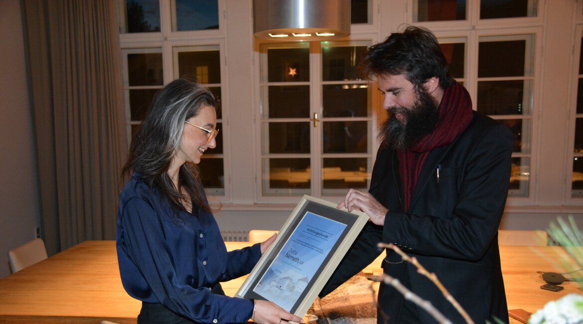Kulturreferent Klaus Reitberger überreicht Zsófia Németh den Kufstein Thesis Award 2024. Sie hält die gerahmte Urkunde mit beiden Händen und lächelt. Im Hintergrund ist ein festlich beleuchteter Raum mit einem großen Holztisch und Fenstern zu sehen.