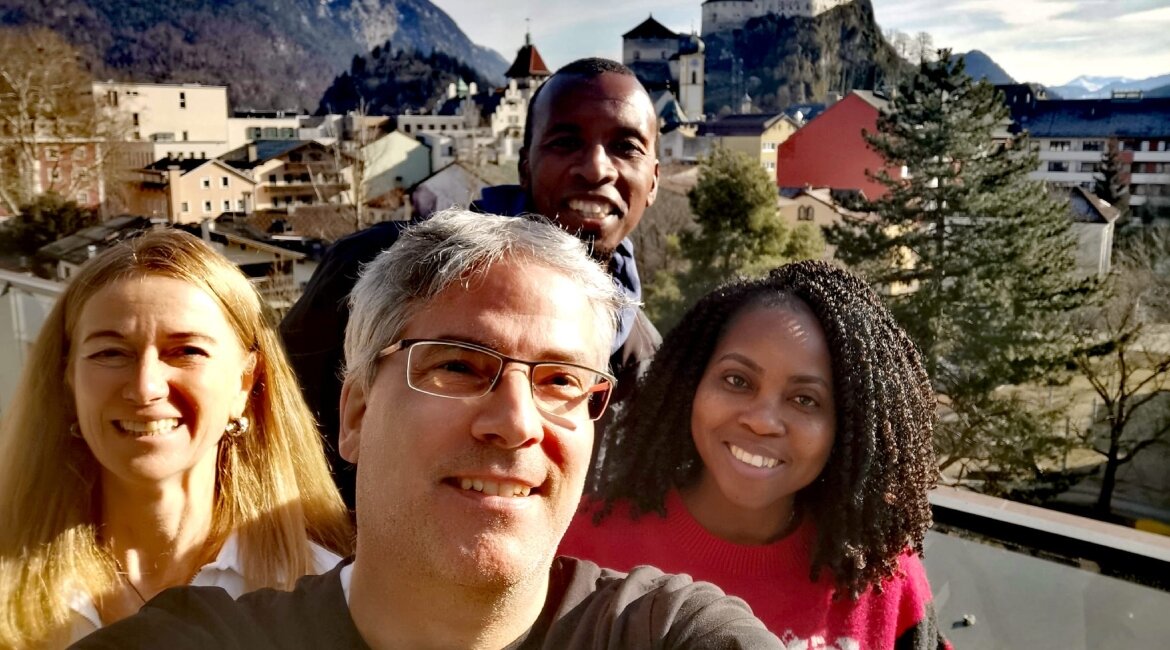 Gruppen-Selfie mit vier Personen auf der Terrasse der HOK. Mag. Uta Fessler-Purkarthofer, MBA (links) und Prof. (FH) Dr. Peter Schneckenleitner (Mitte, 2. von links) von der HOK gemeinsam mit zwei Forschungspartnern aus Mosambik. Im Hintergrund ist die Festung Kufstein zu sehen.