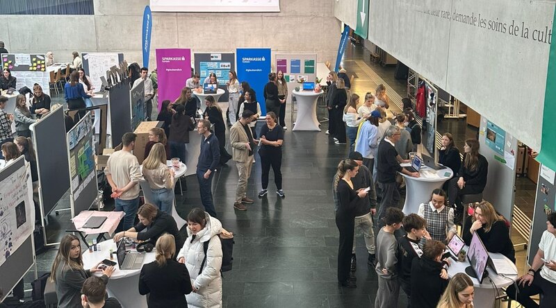 Blick von oben auf die Aula, wo mehrere Besucher:innen und Stände der Expo 2025 der HOK zu sehen sind. 