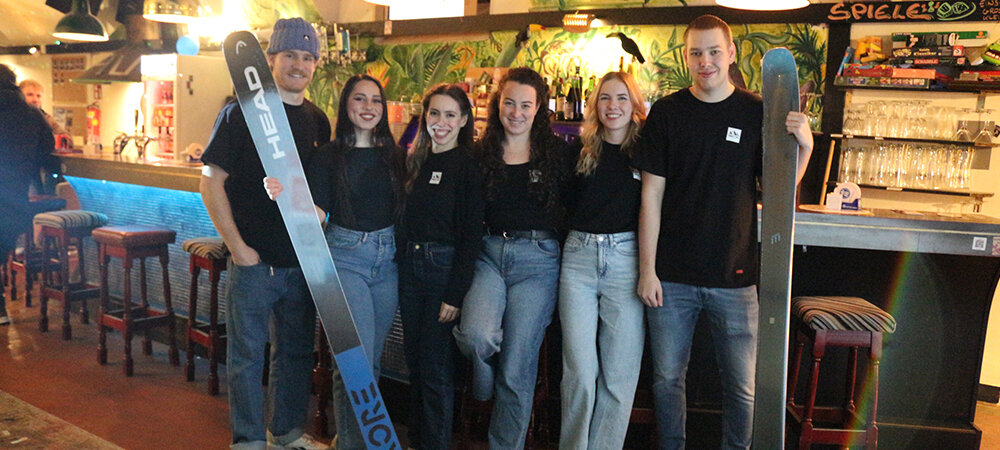 6 Personen stehen in Jeans und schwarzen T-Shirts vor einer Bar. Zwei haben jeweils einen Ski in der Hand.