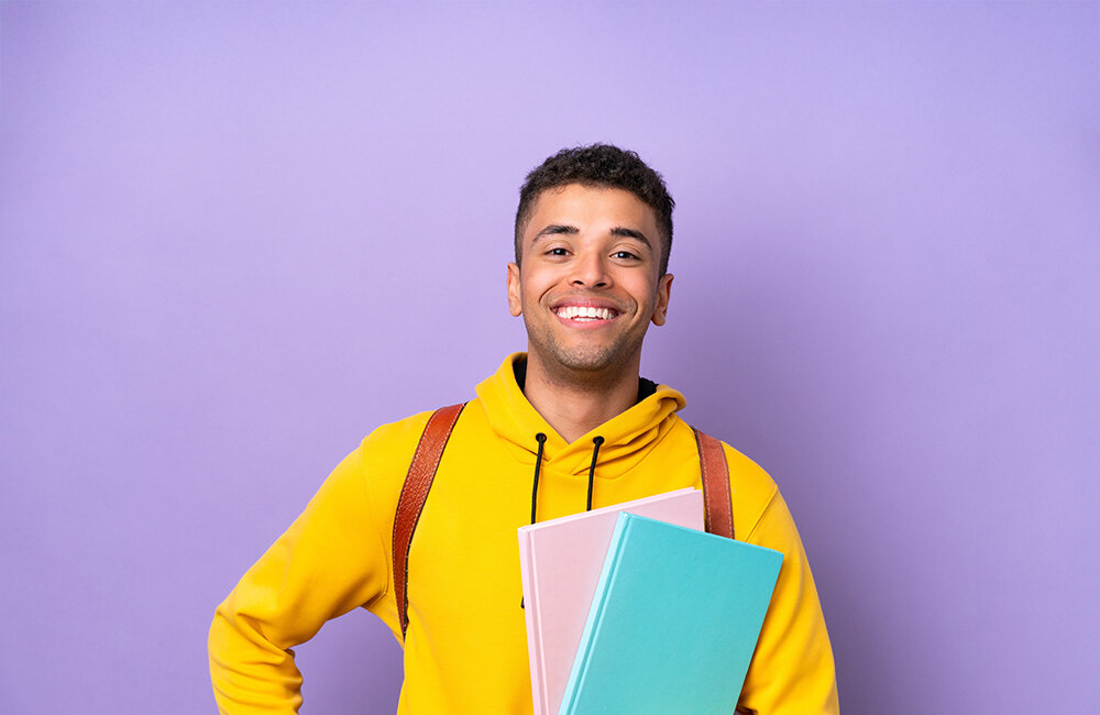 Junger Mann mit Büchern in der Hand und gelbem Pulli vor fliederfarbenen Hintergrund. 