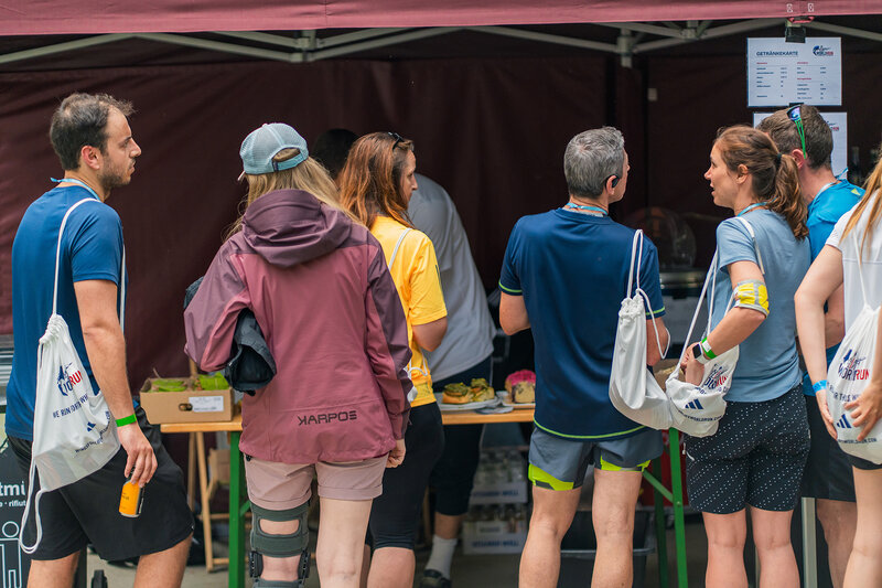Teilnehmende stehen am Verpflegungsstand mit dem Rücken zur Kamera