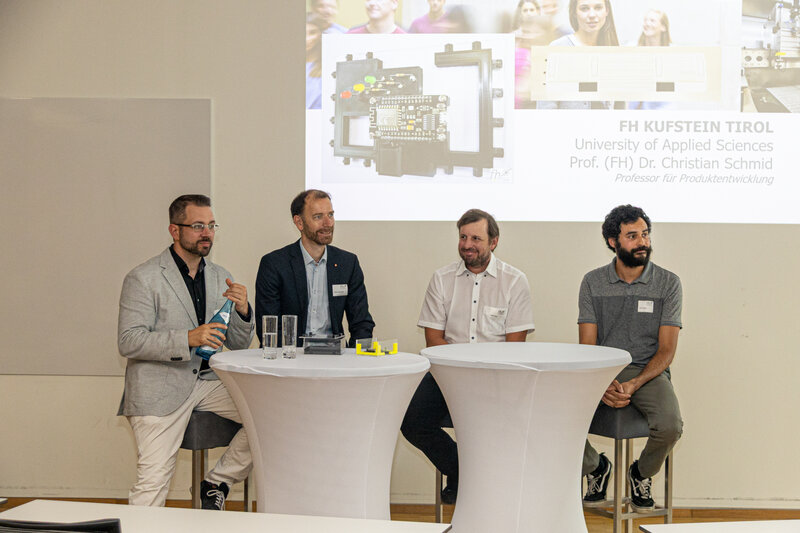 Vier Personen sitzen vor einem Stehtisch, im Hintergrund eine Präsentation.