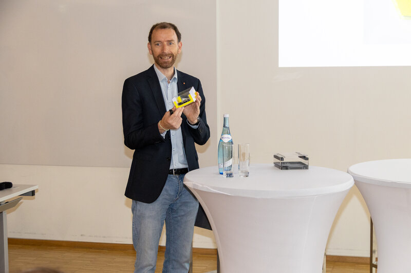 Ein Mann hat einen gelben Zettel in der Hand und steht vor einem Stehtisch.