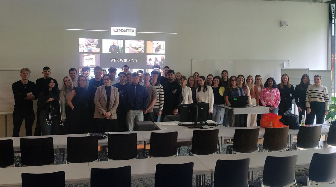 EIne Gruppe Menschen steht in einem Klassenraum vor einer Präsentation.
