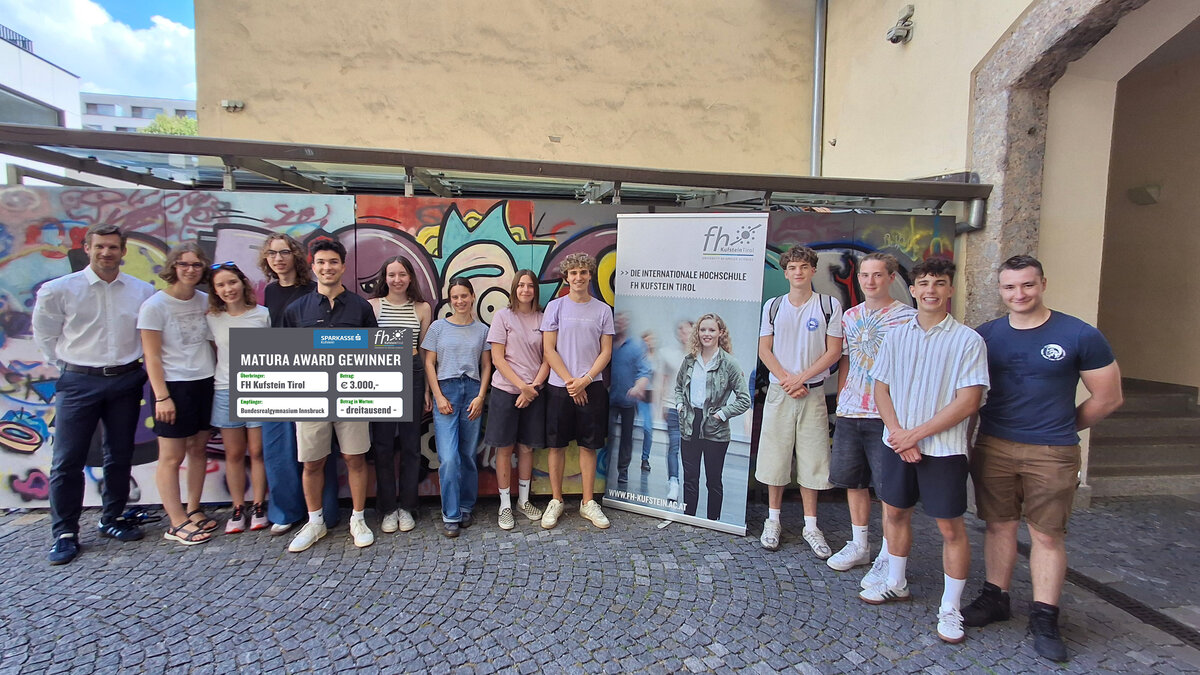 Schüler der 8a des BRG Adolf-Pichler-Platz posieren mit dem Scheck des diesjährigen Matura-Awards vor einer Wand mit Graffiti. 