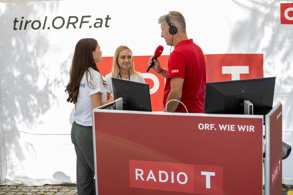 ORF-Moderator im roten Poloshirt interviewt zwei Studentinnen im weißen T-Shirt mit der Aufschrift „HOK“ am Radiostand der Sommerfrische 2025.