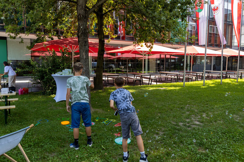 Zwei Kinder spielen im Freien mit Seifenblasen auf der Wiese vor der HOK, während bunte Spielgeräte am Boden liegen.