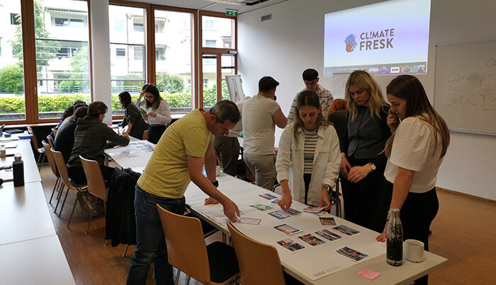 Teilnehmende arbeiten in Gruppen an einem Climate-Fresk-Workshop, Karten liegen auf den Tischen, im Hintergrund läuft eine Präsentation.