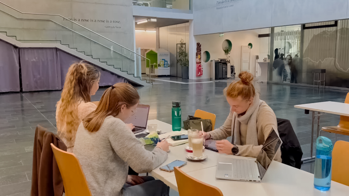 Zwei Studierende und eine Lehrende arbeiten mit Laptops und Unterlagen in der Aula der HOK.