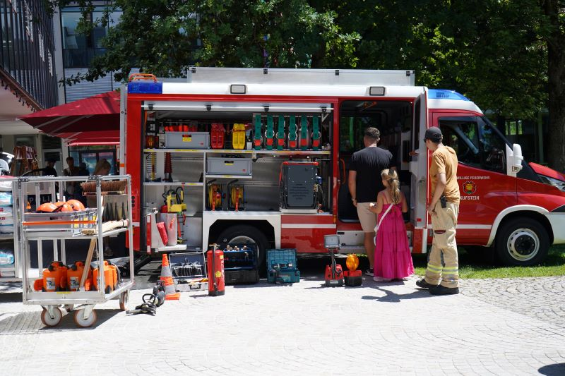 Ein Feuerwehrmann, ein Mann und ein Kind stehen vor einem Feuerwehrauto im Stadtpark vor dem Gebäude der HOK.