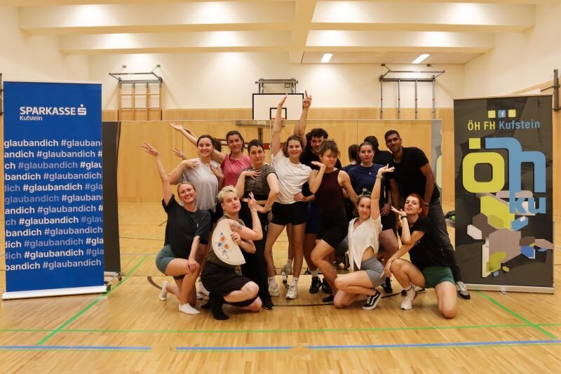 Eine Gruppe von Studierenden posiert in einer Turnhalle nach einem Tanzworkshop des Praxisprojekts Q & U, zwischen Bannern der Sparkasse Kufstein und der ÖH HOK.
