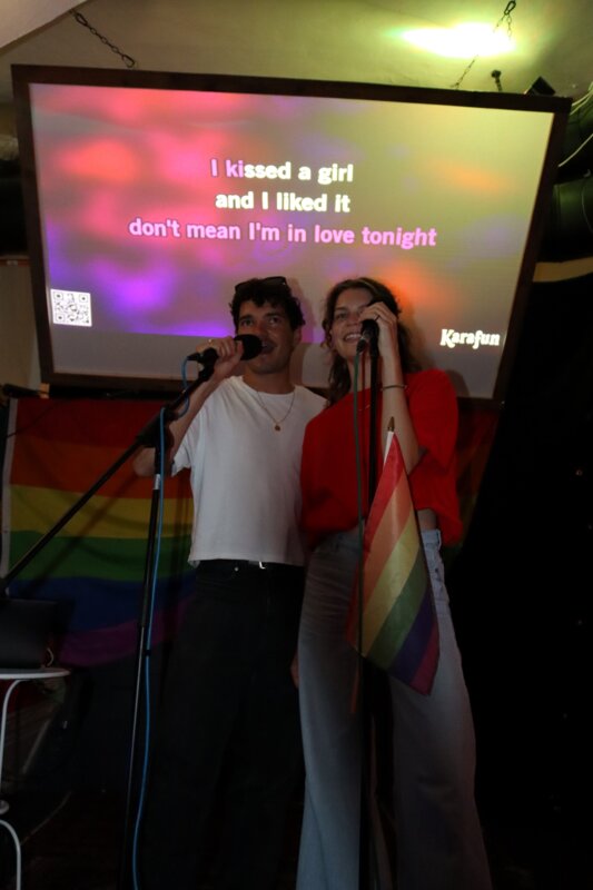 Zwei Studierende singen bei der Karaoke Night des Praxisprojekts QYOU an der HOK. Sie stehen auf einer Bühne vor einer Leinwand mit Liedtext und halten Mikrofone, während eine Regenbogenflagge sichtbar ist.