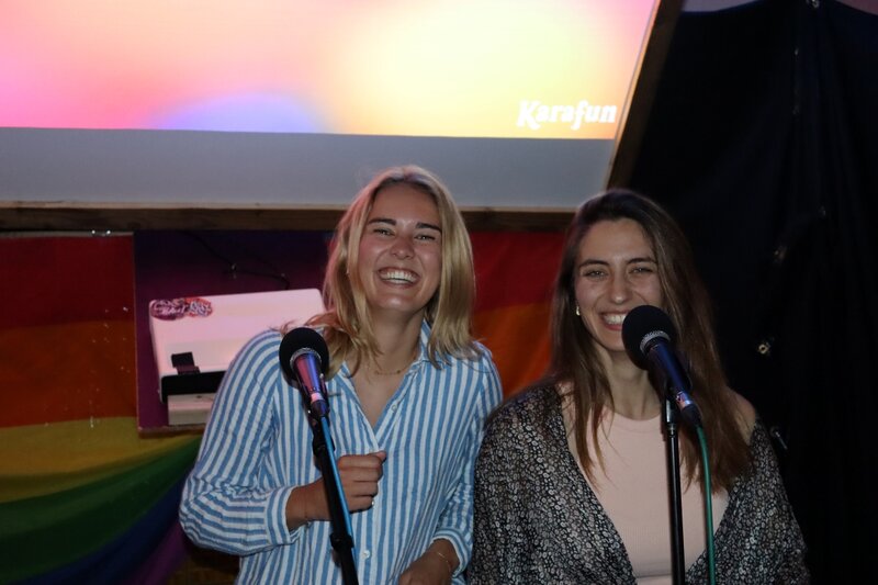 Zwei Studierende stehen lachend auf der Bühne der Karaoke Night des Praxisprojekts QYOU an der HOK. Beide halten Mikrofone in der Hand, im Hintergrund ist eine Leinwand und eine Regenbogenflagge zu sehen.