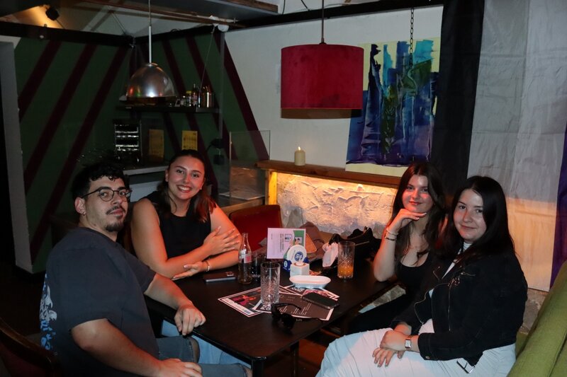 Vier Studierende sitzen an einem Tisch in einer gemütlichen Bar und nehmen am Pub Quiz „Drag Your Brain Out“ im Rahmen des Praxisprojekts QYOU der HOK teil. Getränke und Quiz-Unterlagen liegen auf dem Tisch.